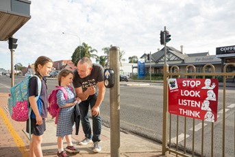 Children and an adult are standing next to a PAC button. The adult is pointing at the button. A safety sign is also visible.