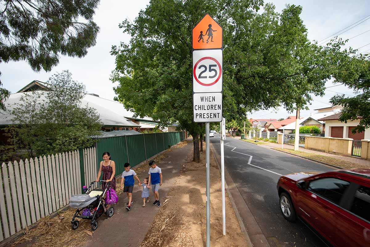 Signs indicating the school zone speed limit are visible. An adult is walking with a pram and two children on the footpath.