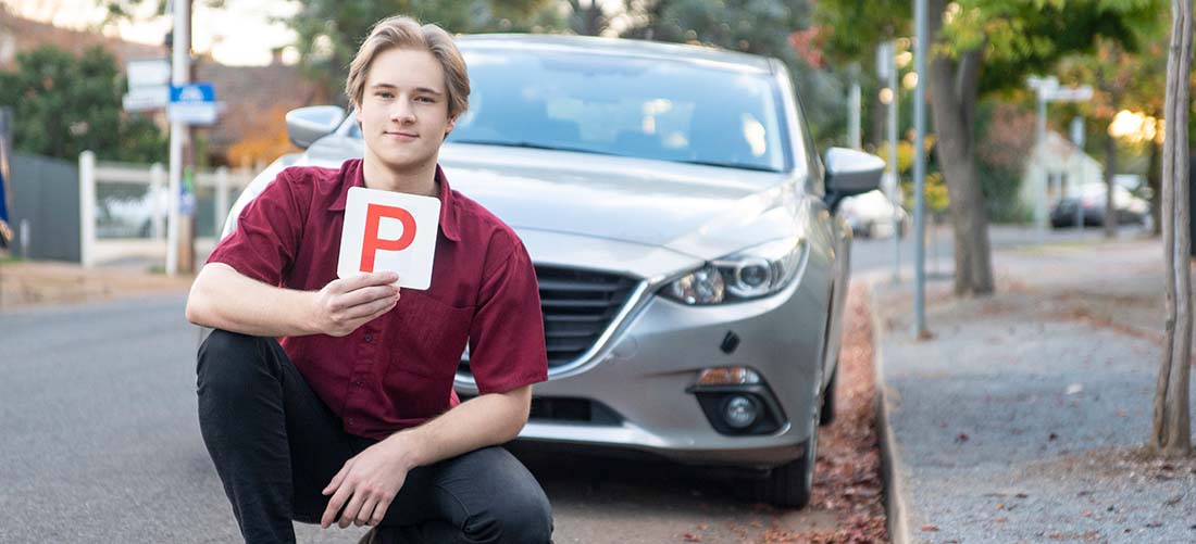 A young man kneeling in front of his car on the road holding a P plate sign in his right hand 