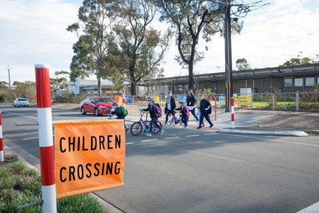 A bright orange "Children crossing" flag is in the foreground while children and adults cross the road in the background.